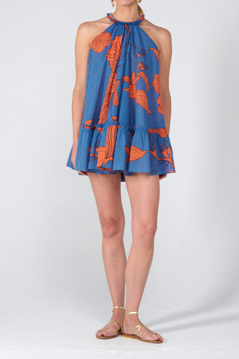 A woman modeling a sleeveless halter-neck mini dress with a blue base and orange tropical floral print, paired with gold-toned flat sandals, against a light gray studio background.