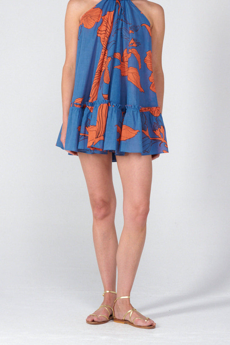 A woman modeling a sleeveless halter-neck mini dress with a blue base and orange tropical floral print, paired with gold-toned flat sandals, against a light gray studio background.