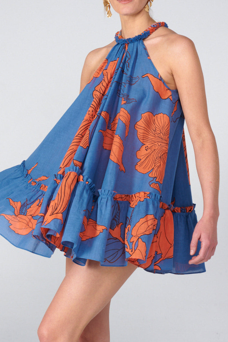 A woman modeling a sleeveless halter mini dress with a blue base and orange floral print, photographed against a light gray studio background.
