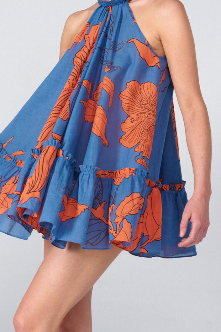A woman modeling a sleeveless halter mini dress with a blue base and orange floral print, photographed against a light gray studio background.