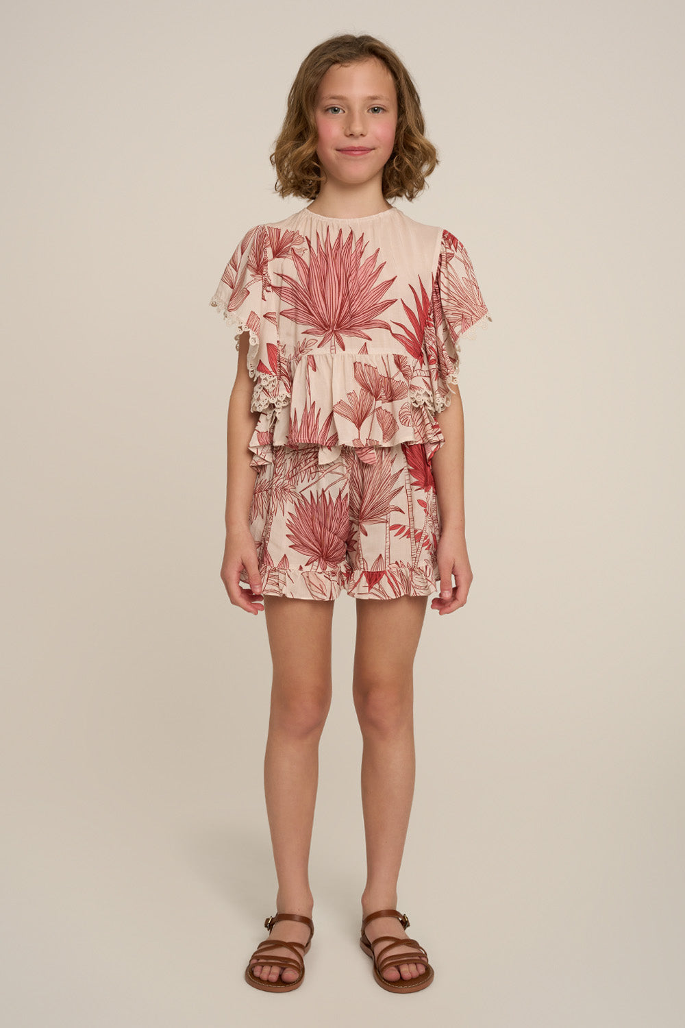 A young girl stands against a plain beige studio background wearing a short-sleeve ruffled romper with a pink and red botanical leaf print on a light peach base, paired with brown strappy sandals.