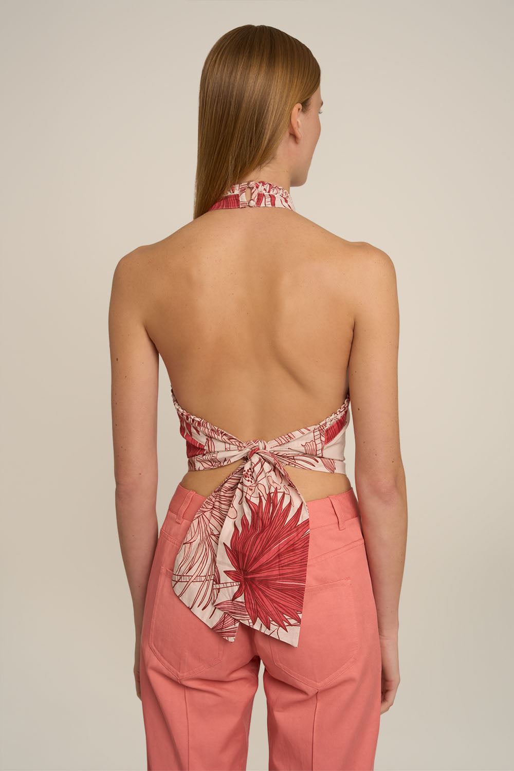 A woman shown from the back wearing a coral pink and red tropical leaf print halter top with a tie-back detail, paired with matching coral pants, against a plain light beige background.