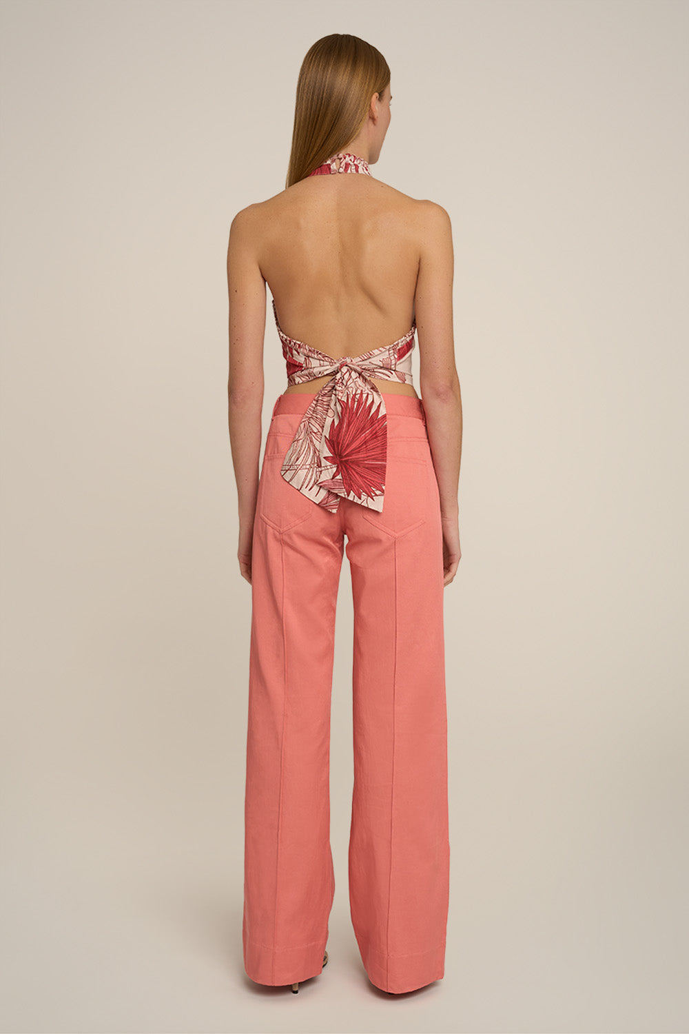 A woman viewed from the back wearing a coral pink halter top with a floral print and tie-back detail, paired with matching wide-leg pants, against a plain beige studio background.
