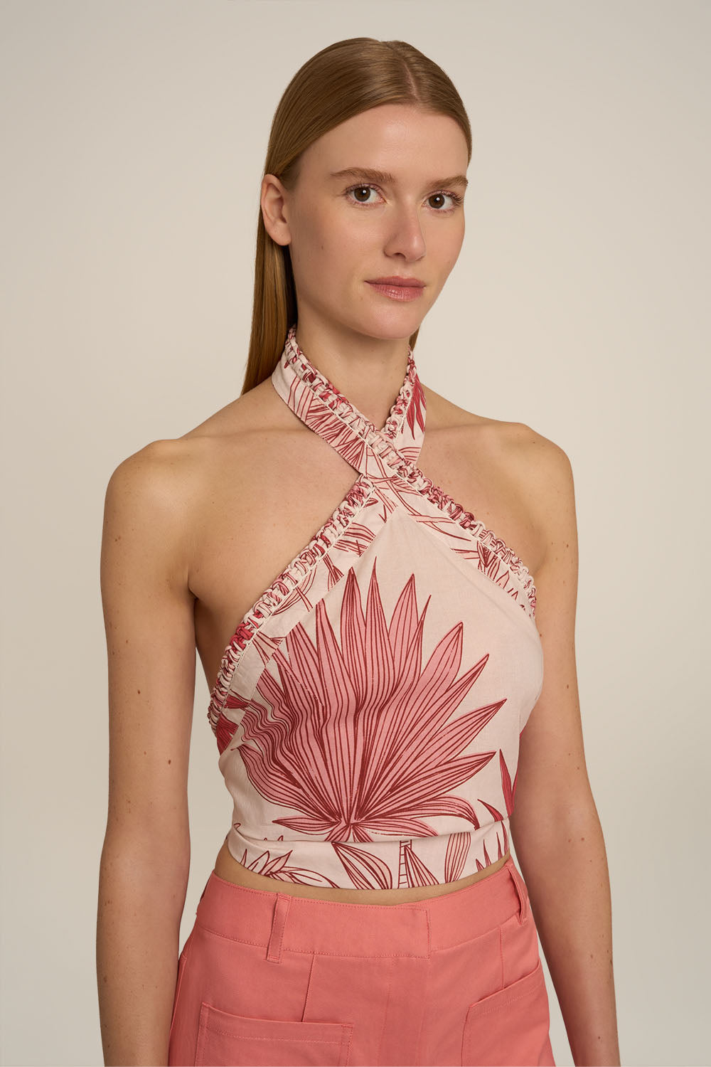 A woman modeling a halter-neck crop top with a pink and beige palm leaf print, paired with pink pants, against a plain beige background.