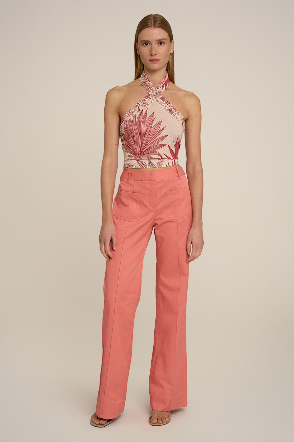 A model stands against a plain beige studio background wearing a sleeveless halter top with a pink tropical leaf print on a light peach base, paired with high-waisted coral pink wide-leg pants.