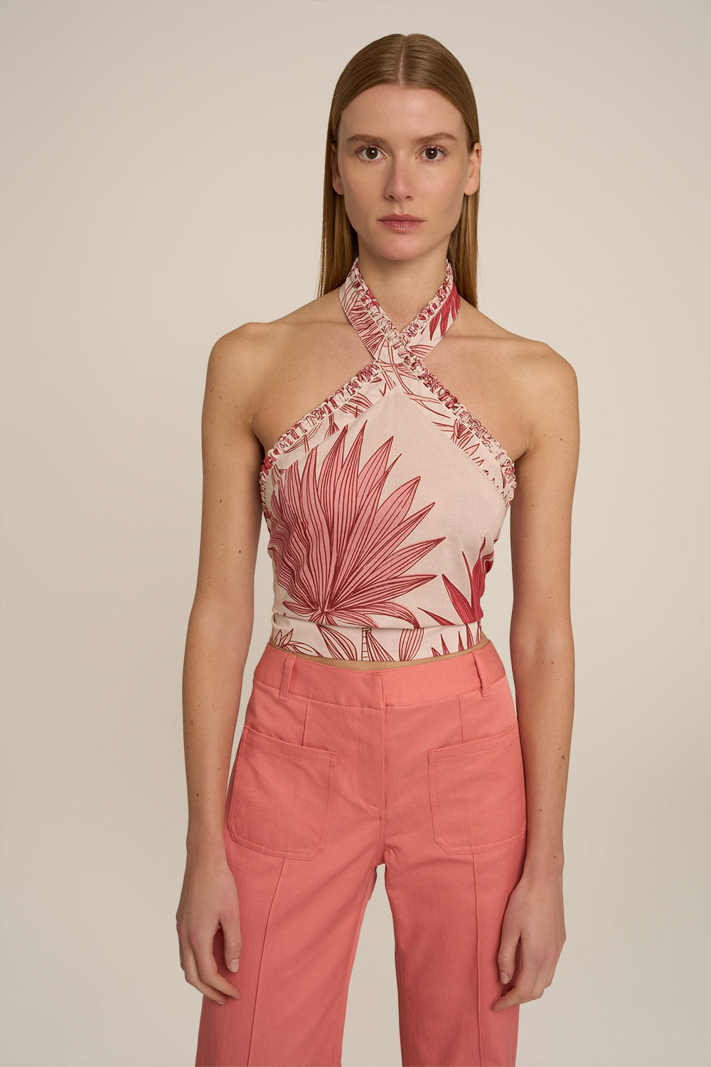 A model wearing a halter-neck crop top with a pink and red palm leaf pattern on a light beige background, paired with high-waisted coral pink trousers, against a solid light beige studio background.
