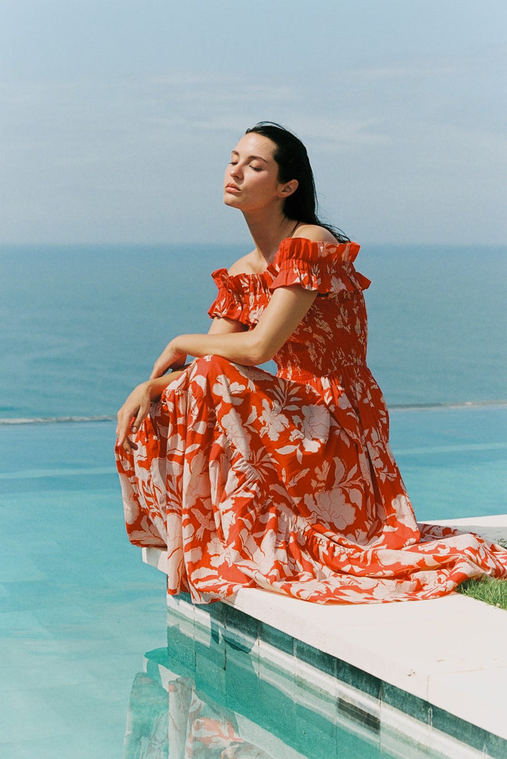 Femme en longue robe orange à imprimé floral assise au bord d'une piscine à débordement, Poupette St Barth