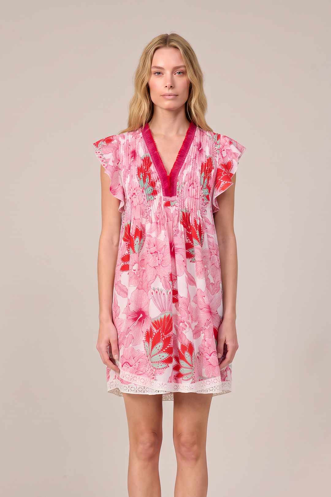 A woman modeling a short pink floral dress with a deep V-neckline, bright pink trim, and flutter sleeves, standing against a solid light beige studio background.